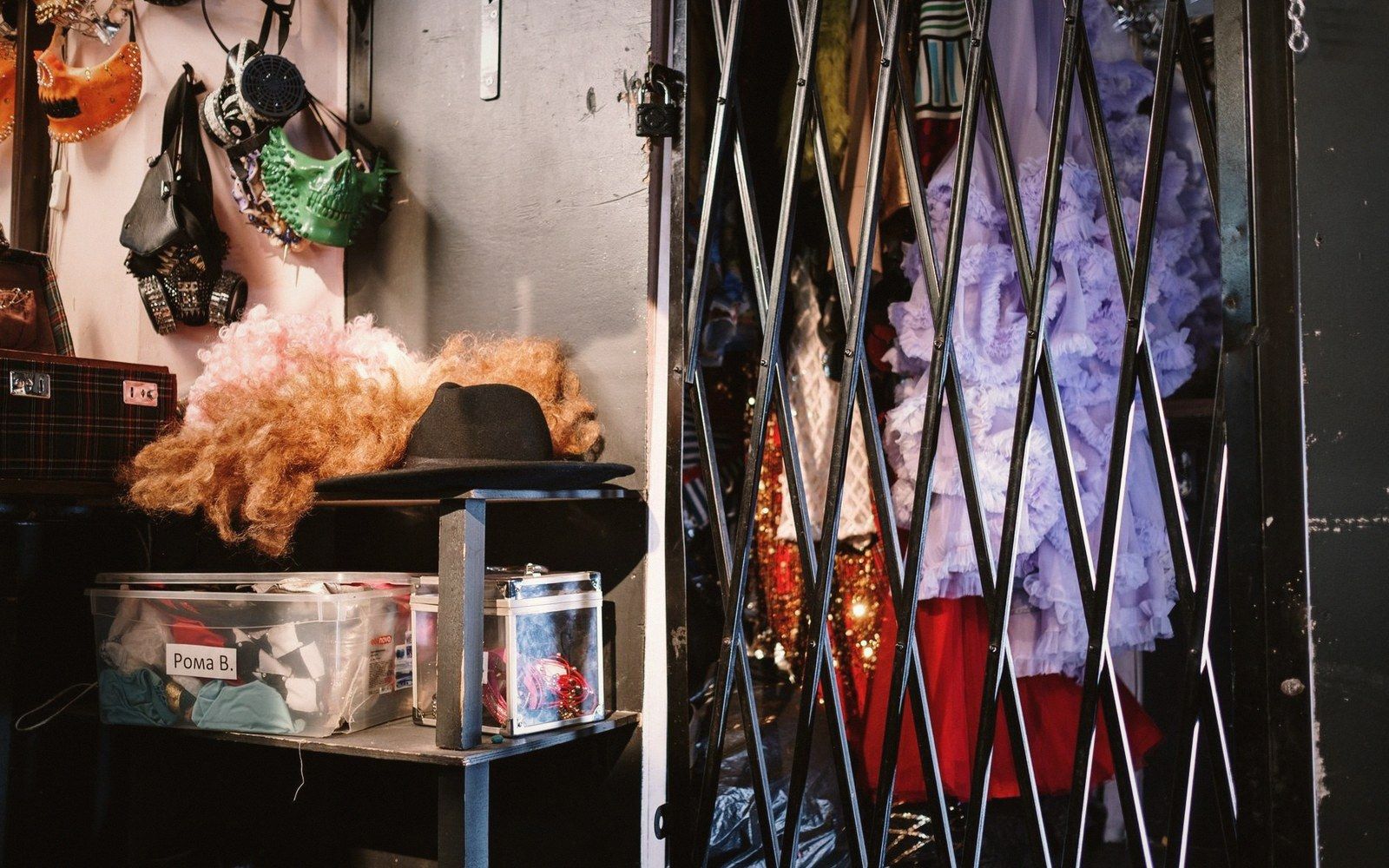 Backstage storage shelves with wigs, masks and hats beside a rack of ruffled costumes behind an accordion gate